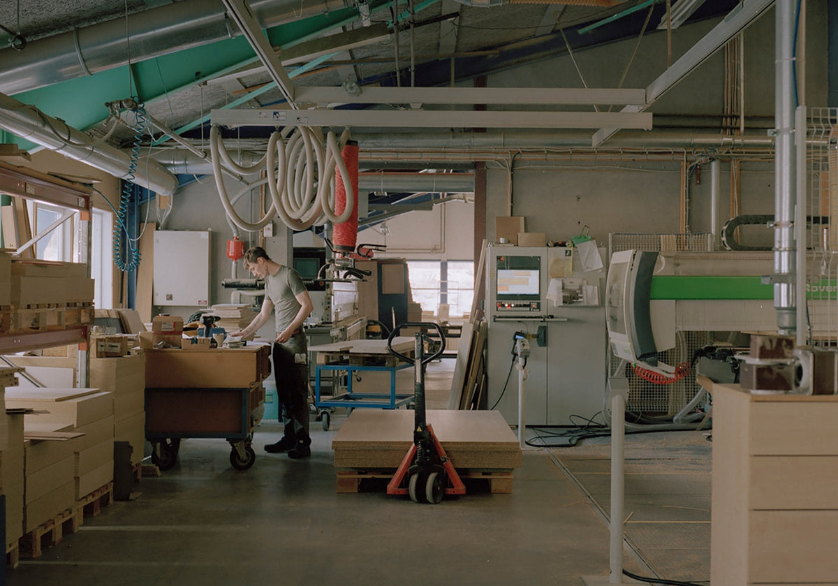 L’atelier Akva : un menuisier à son établi, entouré de panneaux de bois sur palettes, d’un transpalette et d’une machine à commande numérique.