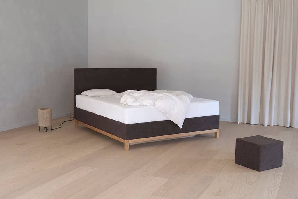 Un lit à eau Carla anthracite à tête de lit droite, posé sur des pieds en bois clair, dans une chambre claire et dépouillée.
