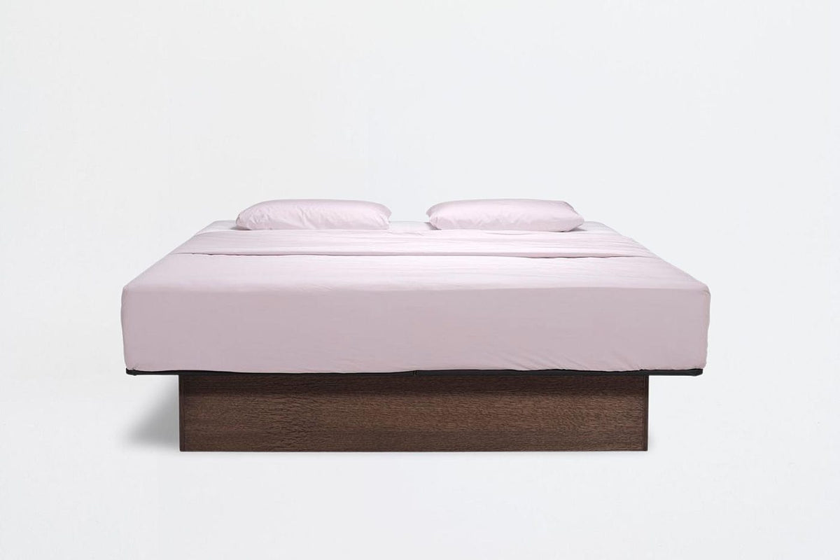 Le lit à eau Soft vu de face sur fond clair : socle en bois foncé, linge rosé et deux oreillers.