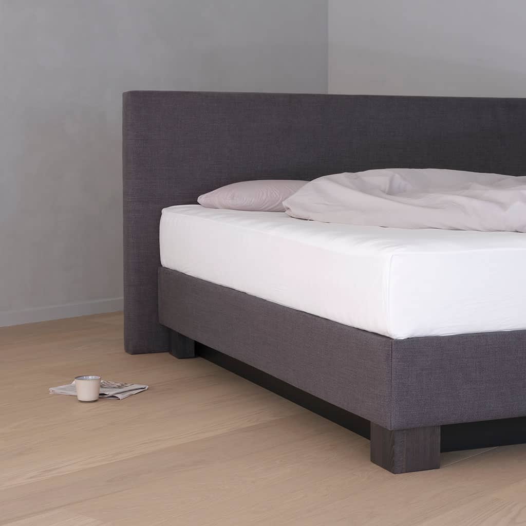 L’angle d’un lit à eau : tête de lit et coffre habillés du même tissu anthracite, posés sur des pieds en bois foncé.
