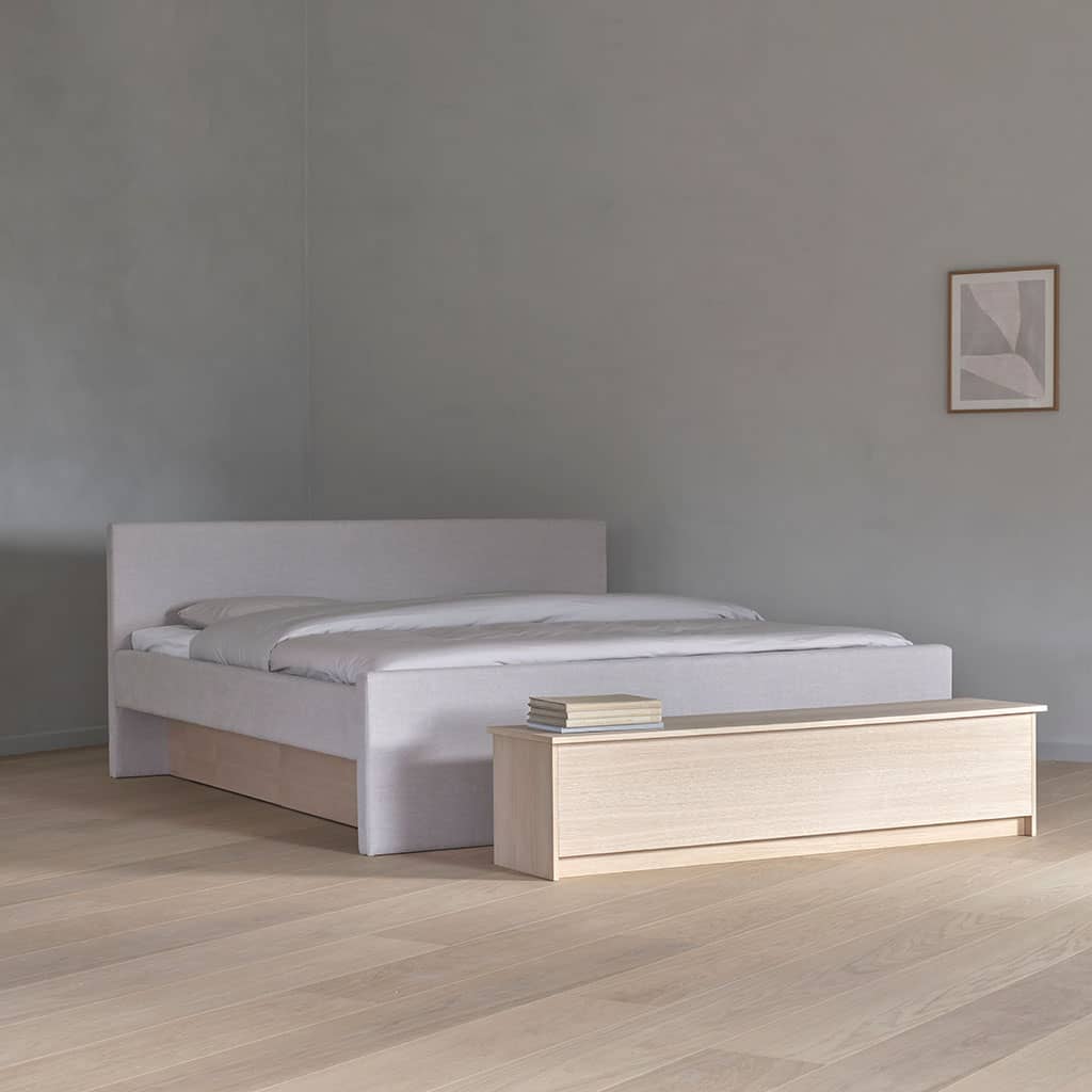 Un lit à eau en tissu clair dans une chambre nue, un banc en bois clair posé au pied du lit.