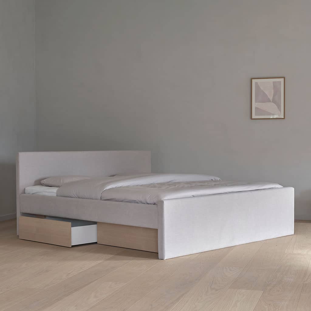 Un lit à eau habillé de tissu clair, tête de lit basse et pied de lit haut, deux tiroirs en bois ouverts sous le socle.