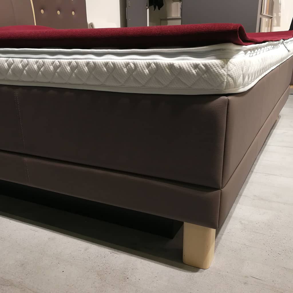 L’angle d’un lit à eau en showroom : le contour habillé de simili-cuir taupe, le revêtement matelassé blanc au-dessus, un pied en bois clair.