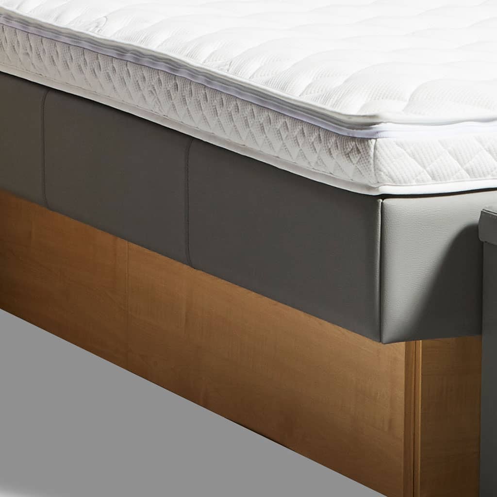 Gros plan sur l’angle d’un lit à eau : le contour en simili-cuir anthracite, le revêtement matelassé blanc, le socle en bois.