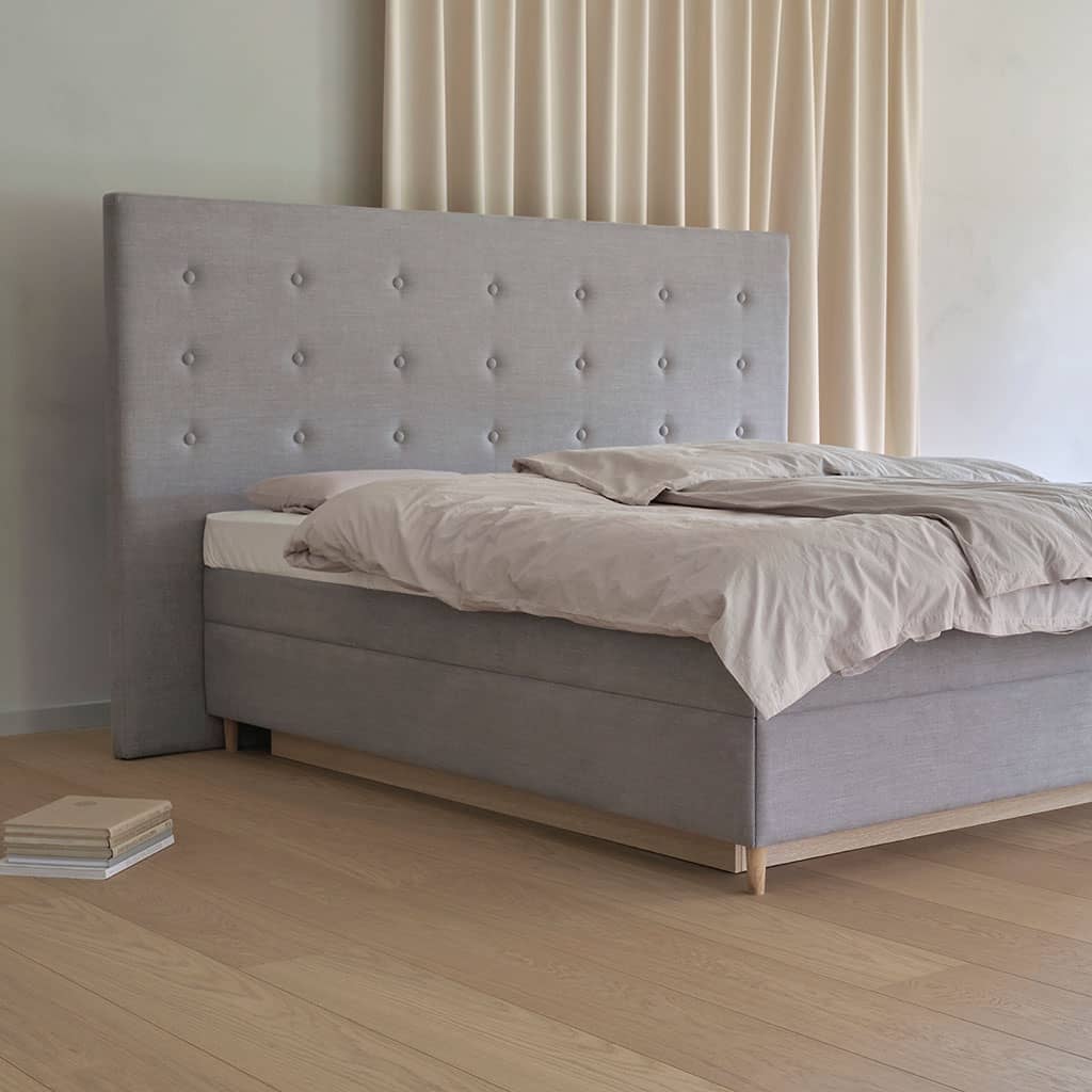Un lit à eau en tissu gris clair avec une tête de lit capitonnée à boutons, posé sur des pieds en bois clair, devant un rideau écru.