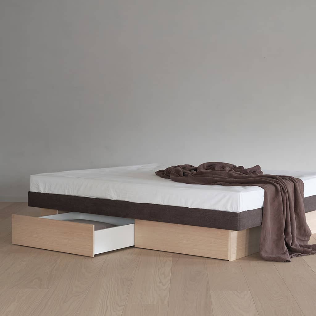 Un lit à eau sans tête de lit, vu de l’angle : socle en chêne clair avec un tiroir ouvert, bandeau sombre du cadre, drap blanc et plaid brun.