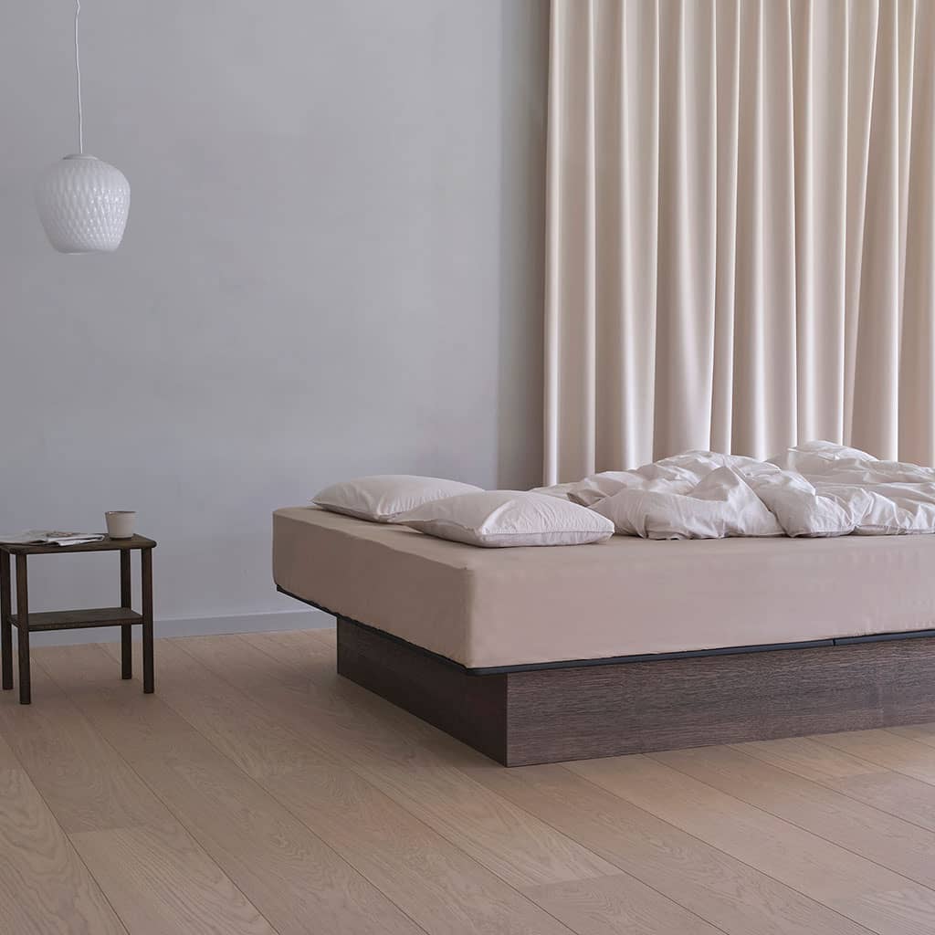 Un lit à eau bas dans une chambre claire : socle en chêne foncé, couchage beige, rideaux écrus et guéridon.
