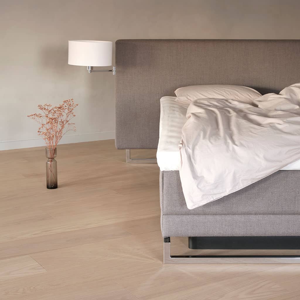 Un lit défait sous une housse de couette terracotta, tête de lit en tissu clair sur montants de chêne, table de nuit et vase.