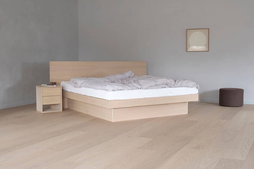 Un lit à eau Nordic en chêne clair à tête de lit pleine, socle bas, dans une chambre claire avec sa table de nuit assortie.