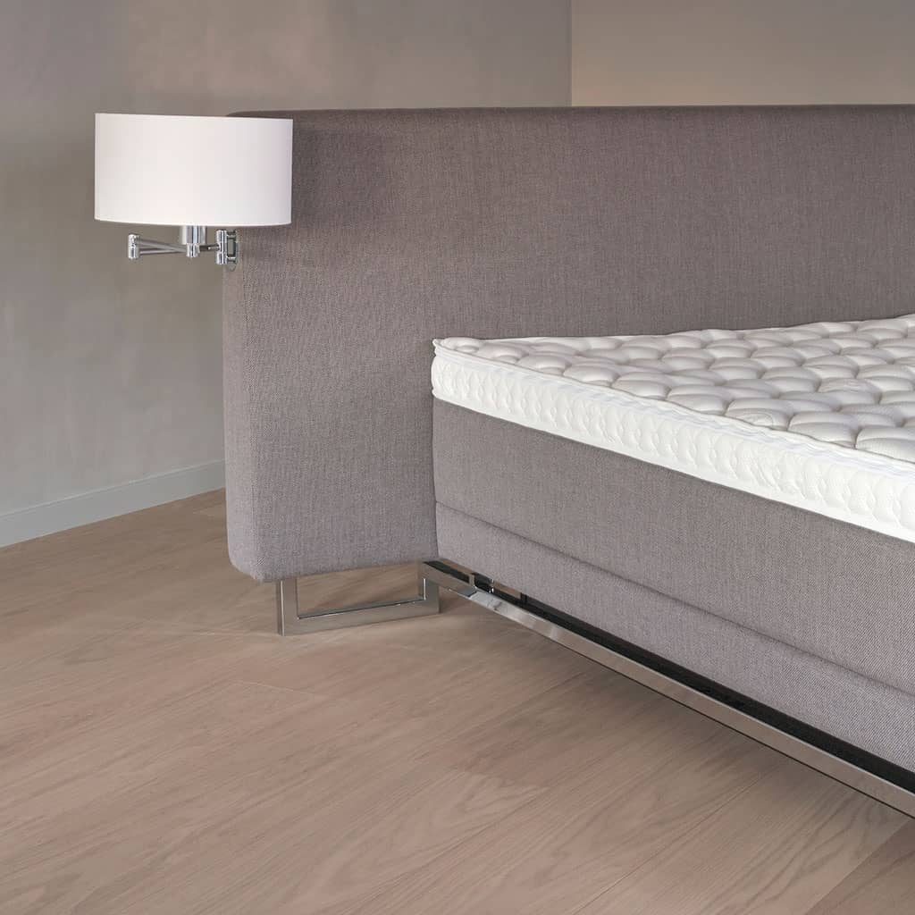 Le lit à eau Vega : tête de lit haute en tissu gris, piètement chromé et surmatelas capitonné, dans une chambre claire.