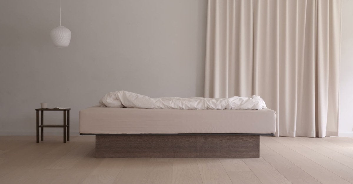 Le lit à eau Akva Soft dans une chambre claire : socle en bois foncé, drap écru et couette blanche, rideaux écrus et petite table de chevet en bois.