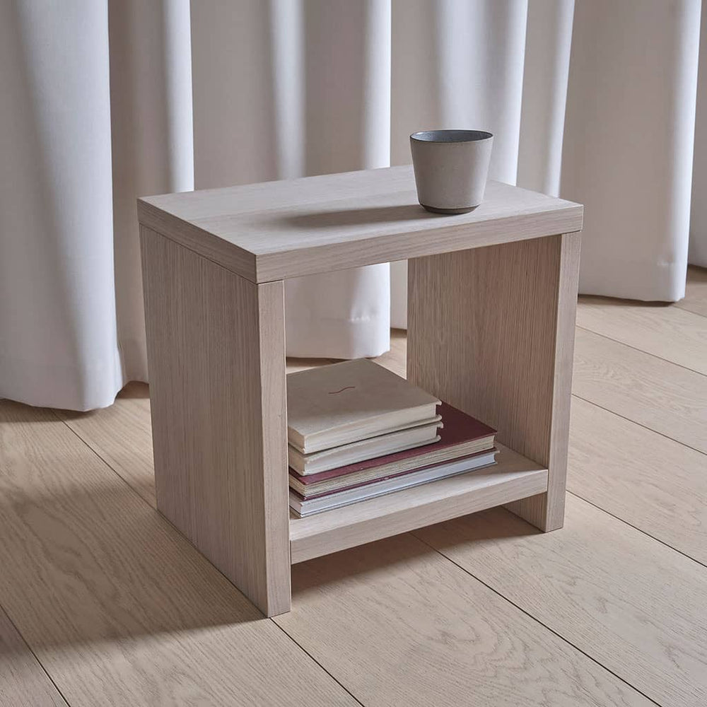 Table de chevet cube Akva en bois clair devant un rideau, livres rangés dans la niche